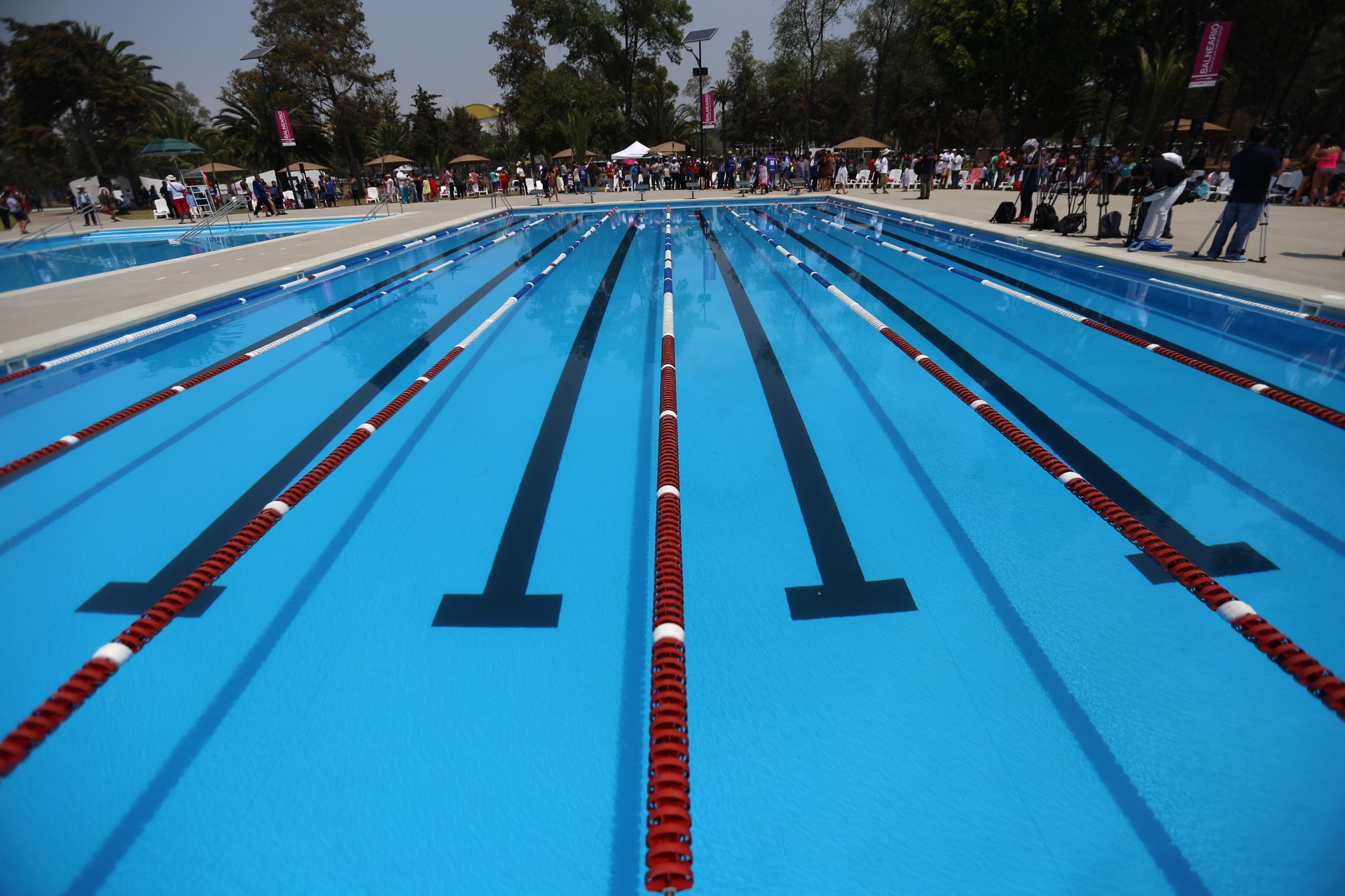 Großer Schwimmbad mit Bahnentrennung, umgeben von Menschen mit Taschen und Gegenständen, sowie Zelten, Bannern, Stangen, einem Schild und einem bewölkten Himmel im Hintergrund.