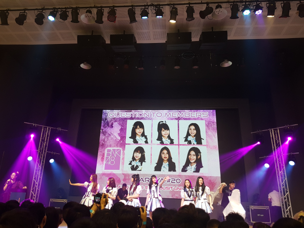 Eine Gruppe von Menschen performs auf einer hell erleuchteten Bühne mit Trägern, während ein Sänger ein Mikrofon hält und ein Musiker ein Instrument spielt, während eine Menge darunter das Ereignis aufzeichnet und ein großer Bildschirm Bilder der K-Pop-Gruppe BK48 anzeigt.