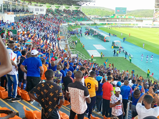 Eine große Menge versammelt sich vor einem eingezäunten Fußballfeld, wobei einige auf Stühlen sitzen und andere stehen, mit Blick auf Hügel unter einem klaren blauen Himmel während der Afrikameisterschaft 2019.