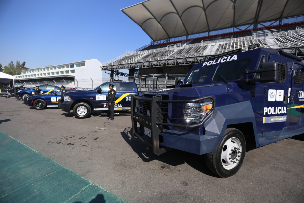 Eine Reihe von Polizeiwagen vor einem Stadion geparkt, mit Menschen im Vordergrund auf der Straße stehend und Gebäuden, Bäumen und einem klaren blauen Himmel im Hintergrund.