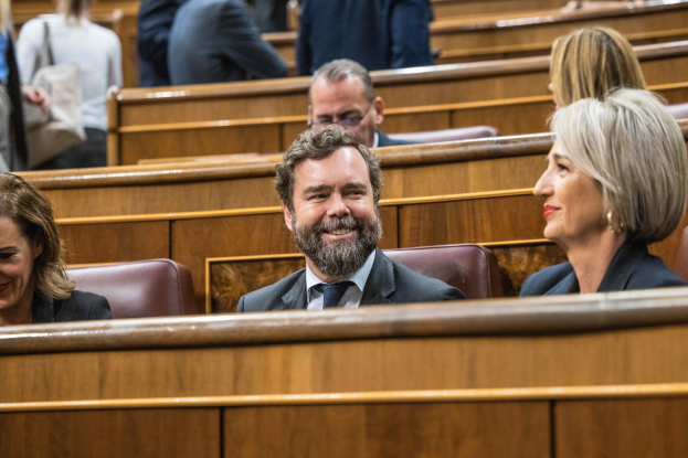 Gruppe von Menschen in einem Gerichtssaal sitzend, mit einem Mann in einem Anzug und zwei Frauen in legerer Kleidung nebeneinander sitzend, Bänke im Hintergrund sichtbar.