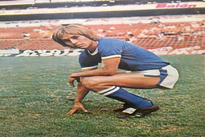 Ein Mann kniet auf einem Fußballfeld, umgeben von sitzenden Personen und Stühlen mit Text darauf, trägt Schuhe.