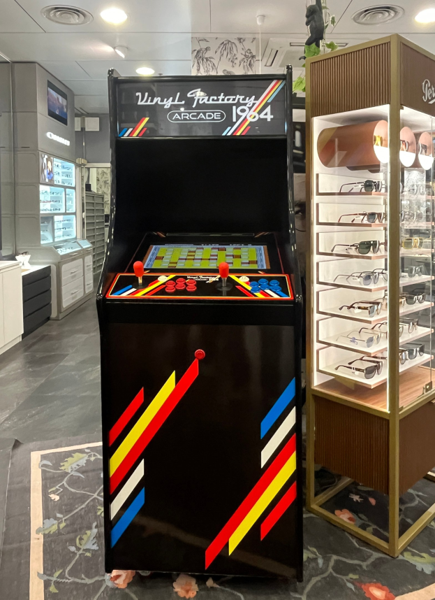 Ein Geschäft mit einem zentralen Retro-Arcade-Automaten, einer Glasvitrine auf der rechten Seite und einer retro-stylischen Beleuchtung mit Schränken und einem Fernseher im Hintergrund.