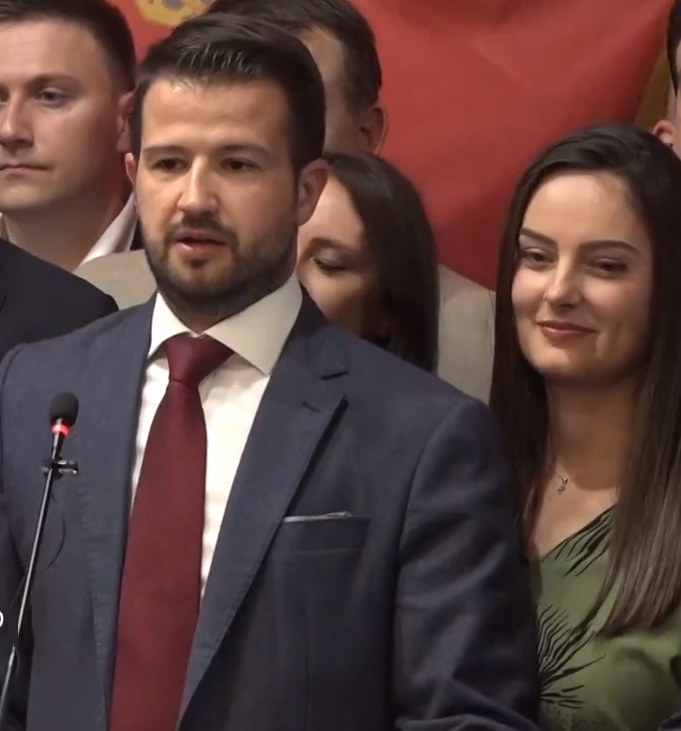 Ein Mann in Anzug und Krawatte spricht in ein Mikrofon an einem Rednerpult, mit einem Banner dahinter und einer aufmerksamen Menge davor.