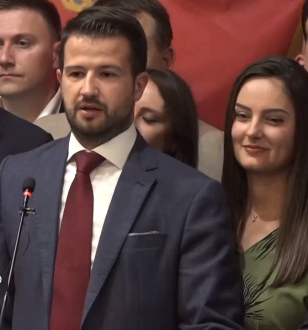 Ein Mann in Anzug und Krawatte spricht in ein Mikrofon an einem Rednerpult, mit einem Banner dahinter und einer aufmerksamen Menge davor.