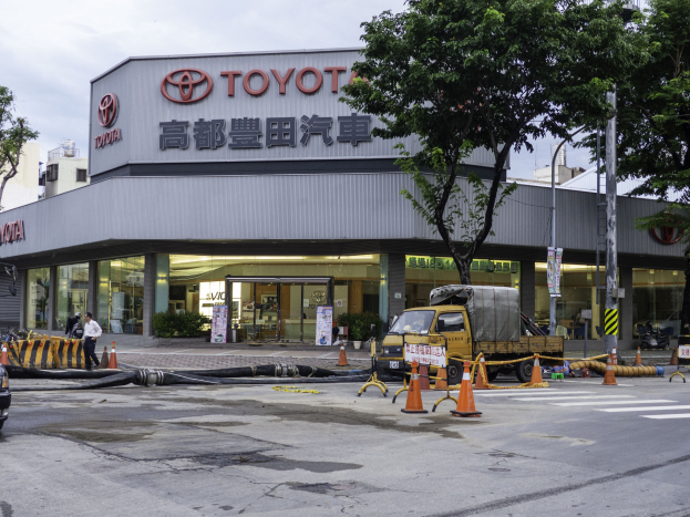 Außenansicht einer Toyota-Vertretung in Tokio, Japan, mit Fahrzeugen, Verkehrskegeln, Rohren, Menschen, Bäumen, Pfählen, Schildern und Gebäuden im Vordergrund unter einem bewölkten Himmel.