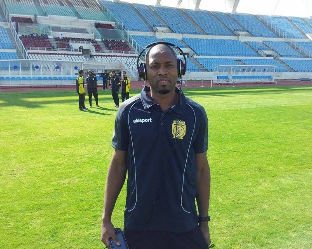 Ein Mann mit Headset, wahrscheinlich der Trainer der ghanaischen Nationalmannschaft, steht auf einem Fussballfeld in der Nähe eines Stadions und hält ein Objekt, während im Hintergrund Menschen zu sehen sind.