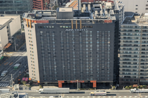 Luftaufnahme des Apa Hotels, eines hohen Gebäudes in einer belebten Stadt in Tokyo, Japan, umgeben von anderen Gebäuden, Fahrzeugen, Pfählen, Bäumen und Pflanzen.