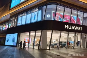 Ein hell erleuchteter Huawei-Store in Shanghai bei Nacht, mit Glaswänden und -türen, mit Menschen, Möbeln und Bildschirmen im Inneren; ein Wasserzeichen erscheint unten.