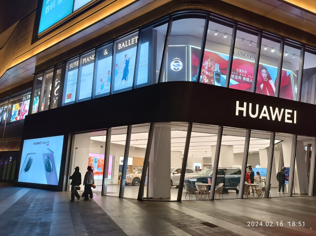 Ein hell erleuchteter Huawei-Store in Shanghai bei Nacht, mit Glaswänden und -türen, mit Menschen, Möbeln und Bildschirmen im Inneren; ein Wasserzeichen erscheint unten.