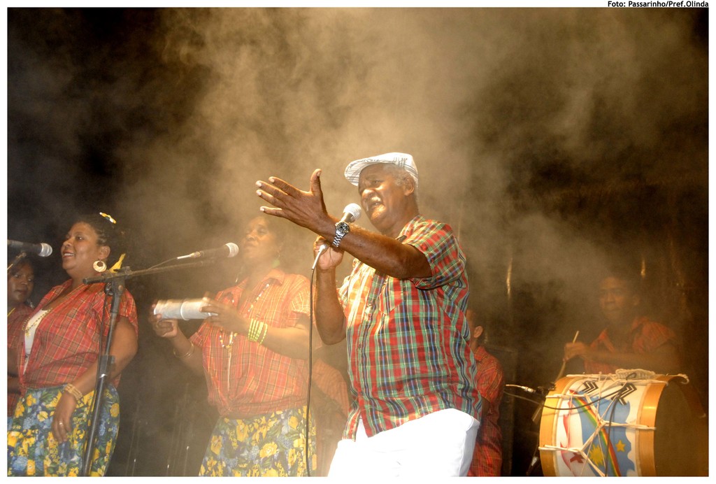 Eine Gruppe von Musikern, die auf der Bühne stehen, mit einem Mann, der in der Mitte in ein Mikrofon singt und einem anderen Mann, der rechts ein Schlagzeug spielt.