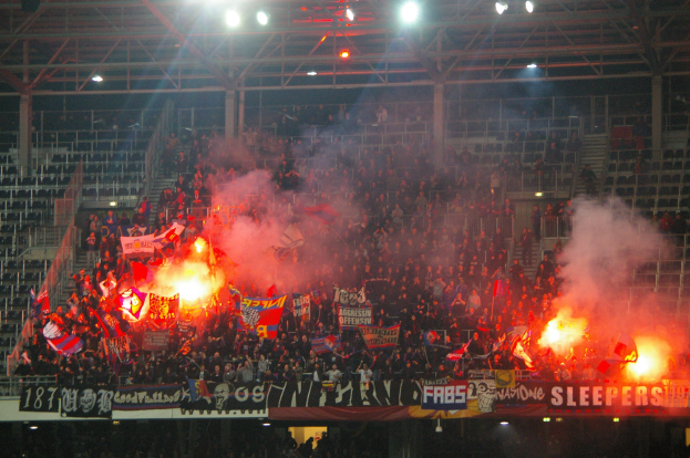 Eine große Menge Menschen in einem Stadion hält Fahnen und Banner, mit Leuchtraketen und Rauch, der aus ihnen aufsteigt, während Metallrahmen und Deckenleuchten über ihnen sichtbar sind.