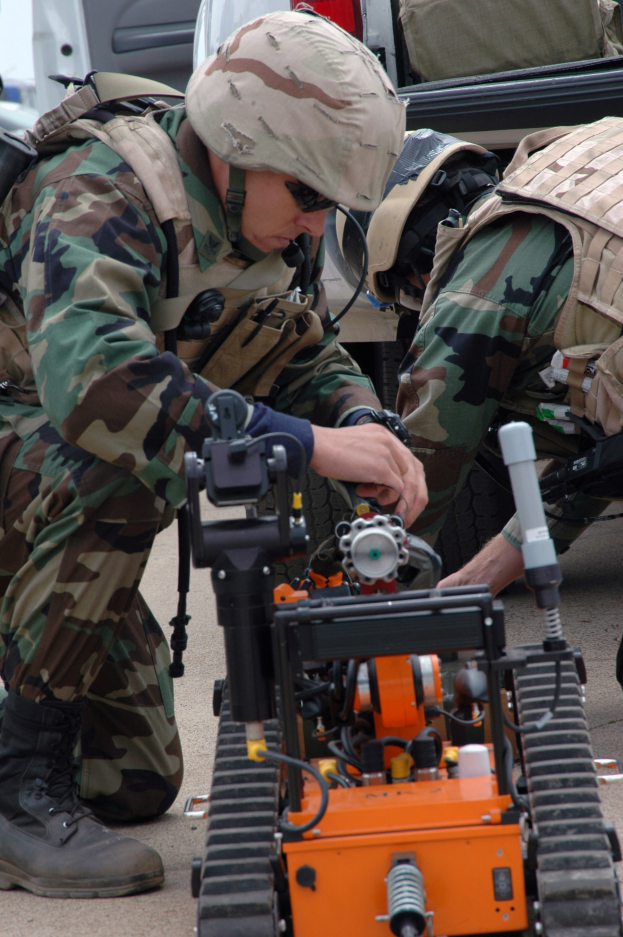 Zwei Männer in Tarnuniformen und Helmen arbeiten an einem kleinen Roboter auf dem Boden, mit Fahrzeugen und Taschen im Hintergrund, was auf eine militärische Umgebung hinweist.