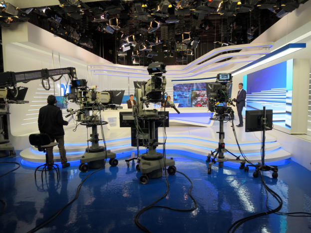 Ein Fernsehstudio mit mehreren Kameras auf dem Boden, einer sitzenden Person im Vordergrund und einigen stehenden Individuen im Hintergrund, mit deckenmontierten Lampen für eine Livesendung.