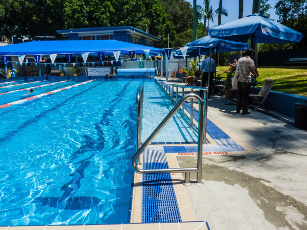 Großer Freibadpool mit mehreren Schwimmern, umgeben von Geländern, Sonnenschirmen, Liegestühlen, Tischen, Grünpflanzen und einem Gebäude unter einem klaren blauen Himmel.