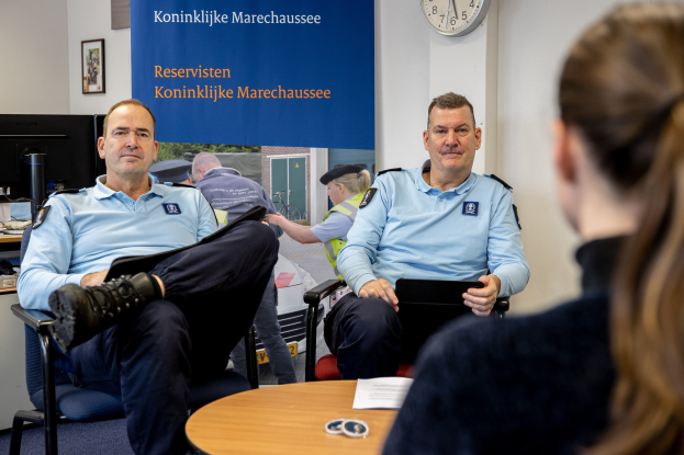 Zwei Polizeibeamte sitzen an einem Tisch und sprechen mit einer Frau; im Hintergrund sind eine Wandtafel, eine Uhr und Foto Rahmen sowie ein weiterer Tisch mit einem Monitor, Tastatur und Maus zu sehen.
