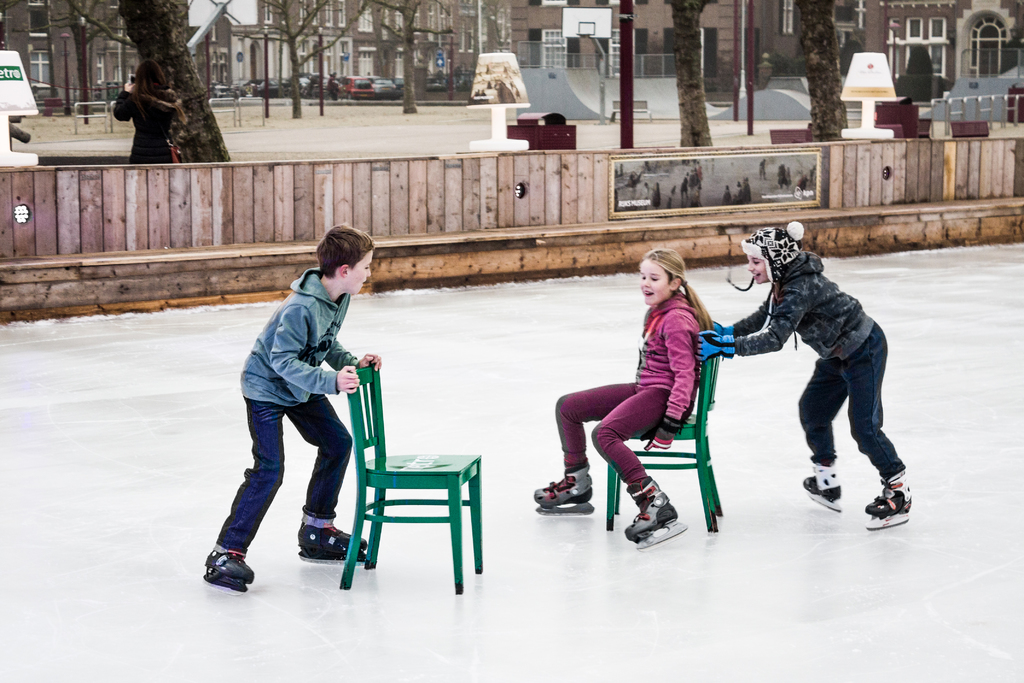 Kinder auf Skiern im Vordergrund eines Spielplatzes mit drei Kindern und zwei Stühlen in der Mitte und Gebäuden, Bäumen, Bänken, Pfosten und einem Basketballfeld im Hintergrund.