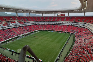 Großes Stadion voller Zuschauer bei einem Fußballspiel mit Hügeln und einem klaren blauen Himmel im Hintergrund.