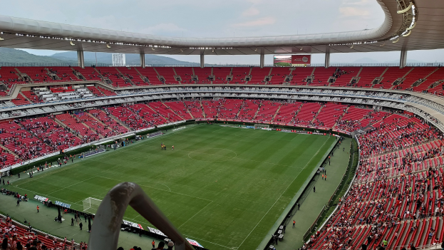 Großes Stadion voller Zuschauer bei einem Fußballspiel mit Hügeln und einem klaren blauen Himmel im Hintergrund.