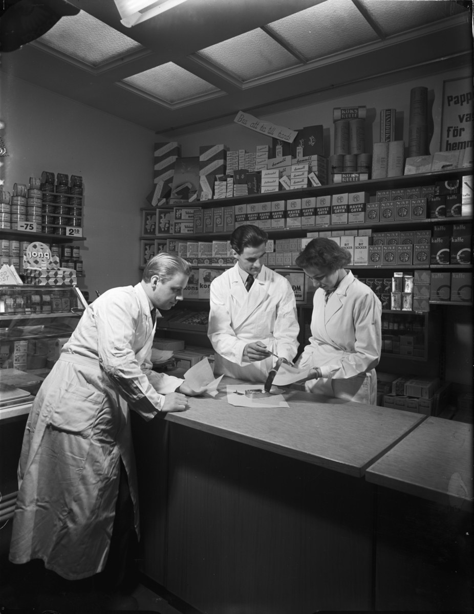 Drei Männer in weißen Kitteln stehen an einem Apothekentresen mit Papieren und Gegenständen, während Regale mit Artikeln im Hintergrund eines Schwarz-Weiß-Bildes zu sehen sind.