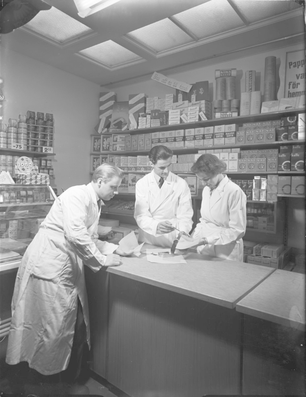 Drei Männer in weißen Kitteln stehen an einem Apothekentresen mit Papieren und Gegenständen, während Regale mit Artikeln im Hintergrund eines Schwarz-Weiß-Bildes zu sehen sind.