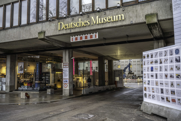 Großes gläsernes Gebäude des Deutschen Museums in Berlin mit einem Namensschild und einer Informationsanzeigetafel auf der rechten Seite, vor dem Hintergrund anderer Gebäude, Bäume und Himmel.