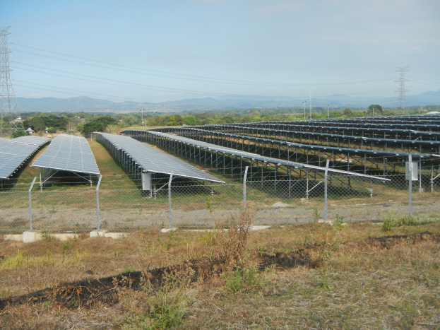 Ein Feld von Solarpanelen hinter einem Zaun, mit Gras und Pflanzen im Vordergrund, Bäumen, Übertragungsmasten mit Drähten und Hügeln im Hintergrund unter einem klaren blauen Himmel.