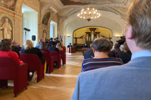 Eine Gruppe von Menschen, die in einem Raum mit Fotorahmen an den Wänden, einer Decke und einem Podium mit Mikrofon und Ständer im Hintergrund sowie Fahnen und anderen Gegenständen an einem Treffen des schwedischen Parlaments teilnehmen.