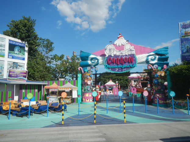 Karussell am Parkeingang mit Straße, Umzäunung, Buden, Banner, Bäumen und bewölktem Himmel im Hintergrund.