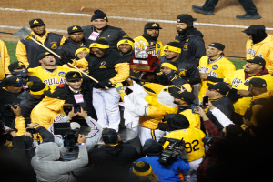 Eine Gruppe von Baseball-Spielern in gelb-schwarzen Uniformen steht auf einem Feld, einige halten einen Pokal, andere halten Kameras und Handys, mit den Beinen einer Person im Hintergrund.