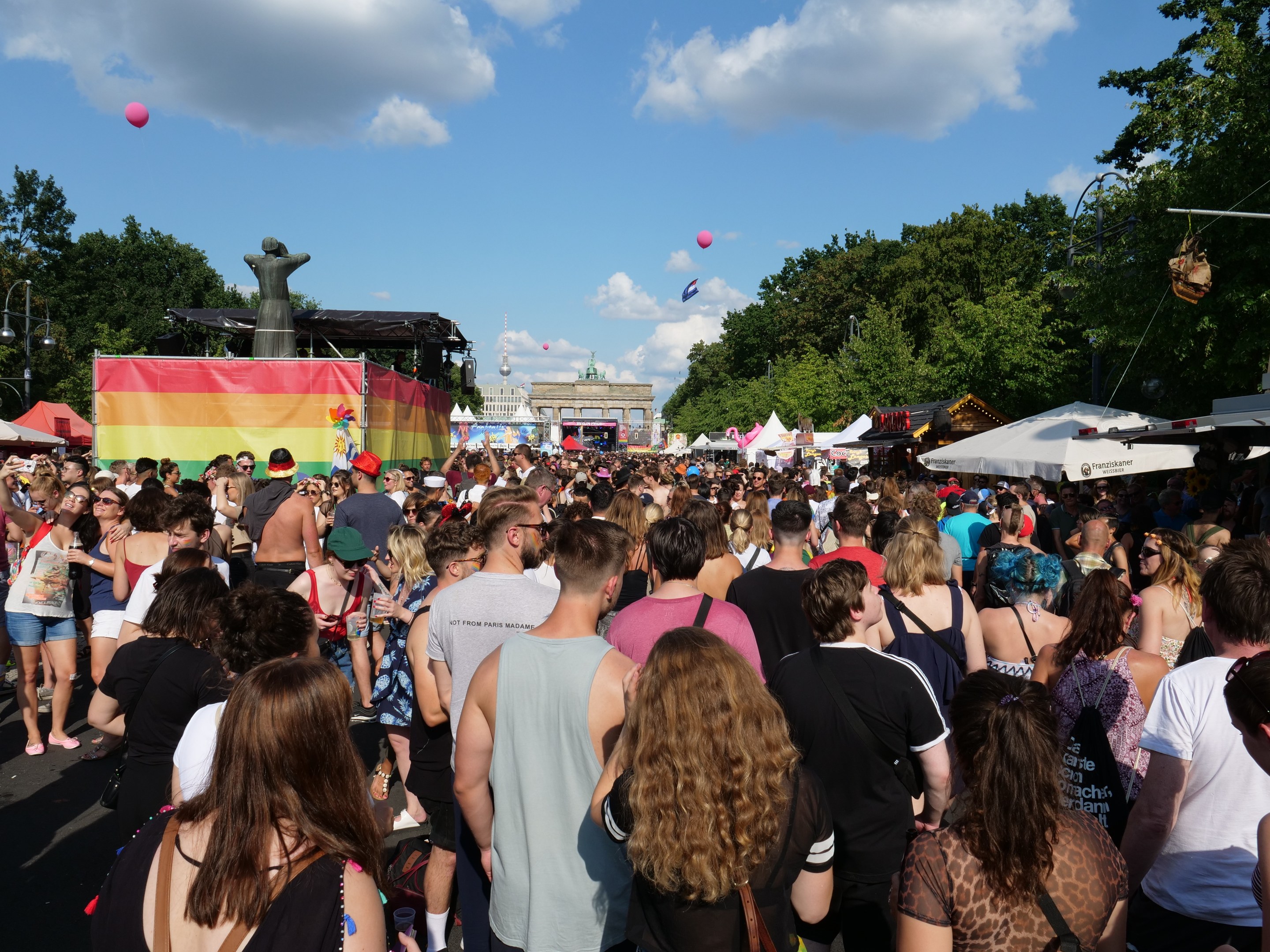 Eine große Menge Menschen marschiert auf einer Straße mit Zelten, Bäumen, Pfählen, Lichtern und einer Statue entlang, im Hintergrund Gebäude und ein Himmel mit Wolken und Ballons bei der Berlin Pride Festival.