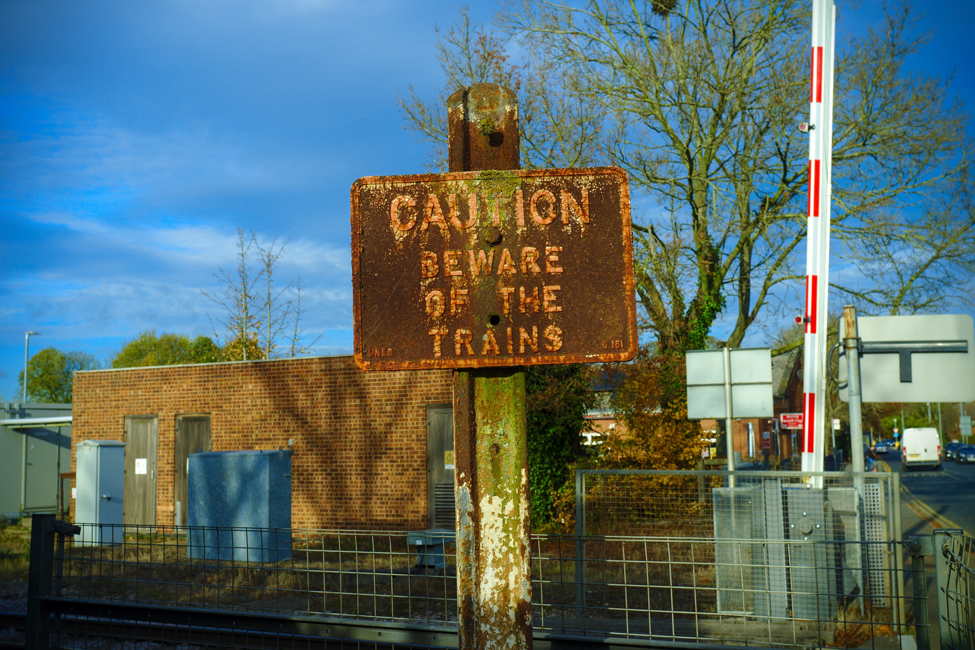 Vorsichtsschild an einem Bahnübergang-Zaun neben Bäumen, Pfählen, einem Gebäude mit Fenstern, Containern, einem Straßenpfahl, Fahrzeugen auf der Straße und einer bewölkten Himmel.