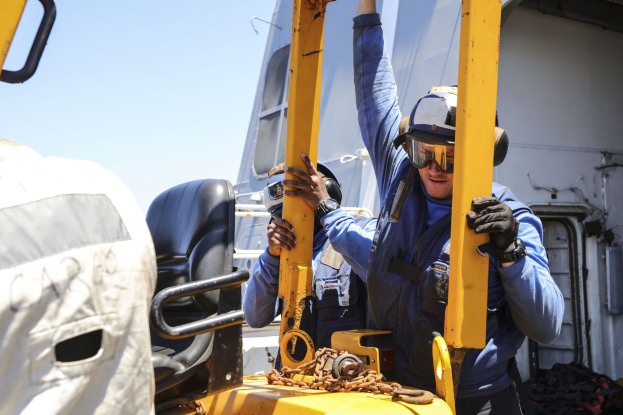 Zwei Männer in blauen Uniformen und Schutzausrüstung arbeiten an einem gelben Gabelstapler, einer hält ein Metallobjekt, im Hintergrund sind ein Fahrzeug und der Himmel zu sehen.