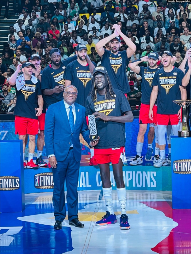 Eine Gruppe von Männern auf einem Basketballfeld, die Trophäen halten und von Zuschauern umgeben sind, mit zwei Trophäen auf Podesten rechts im Bild.