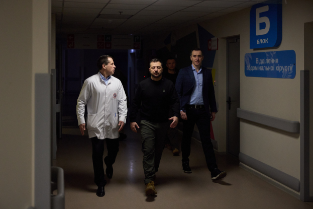 Eine Gruppe von Männern geht einen Krankenhausflur entlang, mit beleuchteter Decke, einer Tür auf der rechten Seite und Texttafeln auf der linken Seite, die besorgt aussehen.
