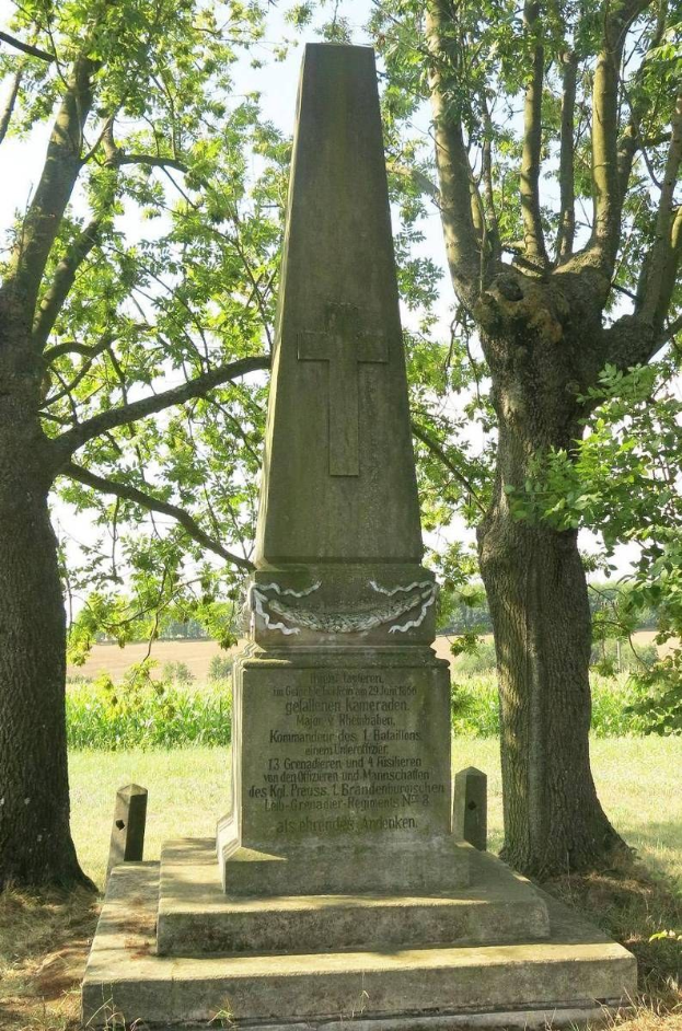 Ein hoher Denkmal mit einem Kreuz steht auf einer grünen Wiese mit Bäumen und einem klaren blauen Himmel im Hintergrund, umgeben von Pflanzen.