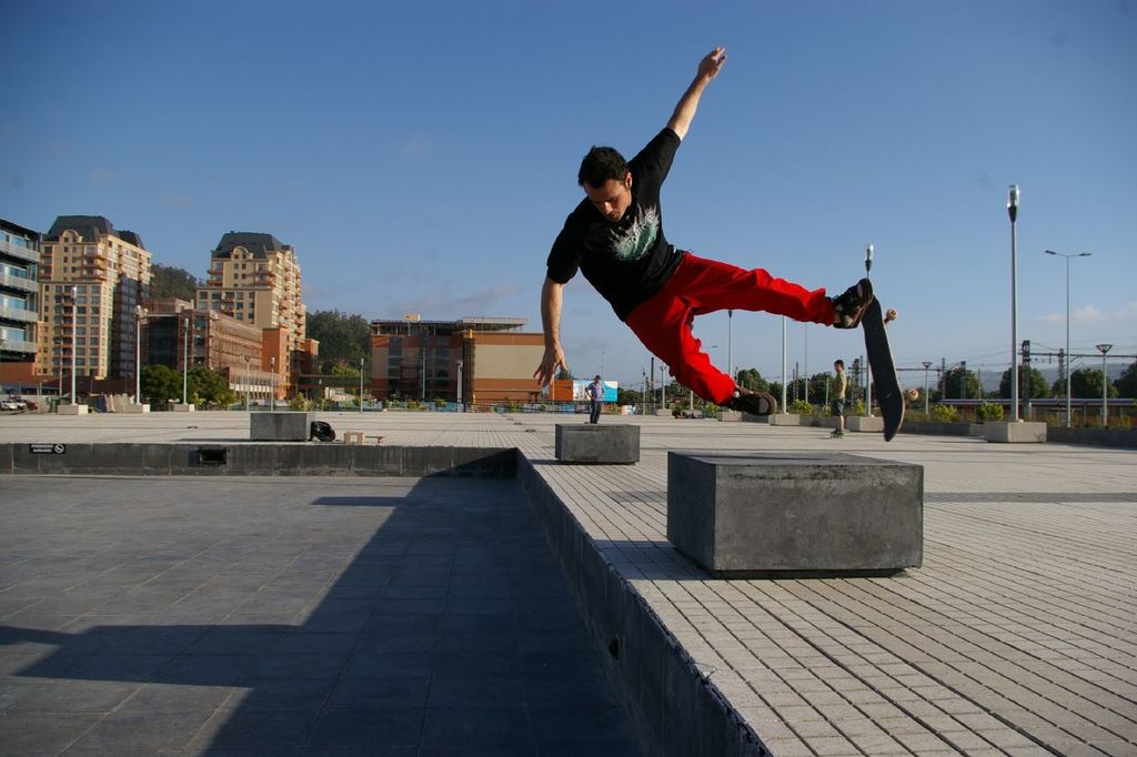 Ein Mann springt auf einem Skateboard mit Gebäuden, Bäumen, Masten und Menschen im Hintergrund und einem Netz auf der rechten Seite.