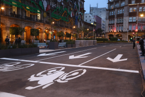 Eine Stadtstraße mit einem Fahrradweg, Gebäude, Straßenlaternen, Verkehrsschilder, Menschen auf dem Gehweg, Bänke, Pflanzen, Bäume, dekorative Gegenstände und einen klaren blauen Himmel.