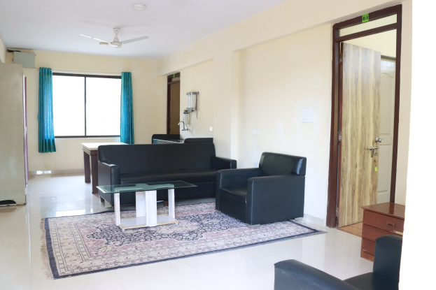 Wohnzimmer mit einer Sitzgruppe, Beistelltischen, einem Teppich, einem wandmontierten Flachbildfernseher, Vorhängen, Deckenleuchten und einer Tür.