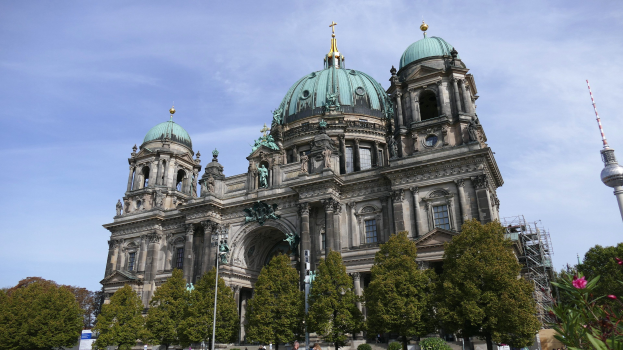 Großes, prunkvolles Berliner Dom-Gebäude in Berlin, Deutschland, mit Statuen, Bögen und Säulen, vor einem bewölkten Himmel, mit Passanten, Bäumen und einem Turm im Vordergrund.