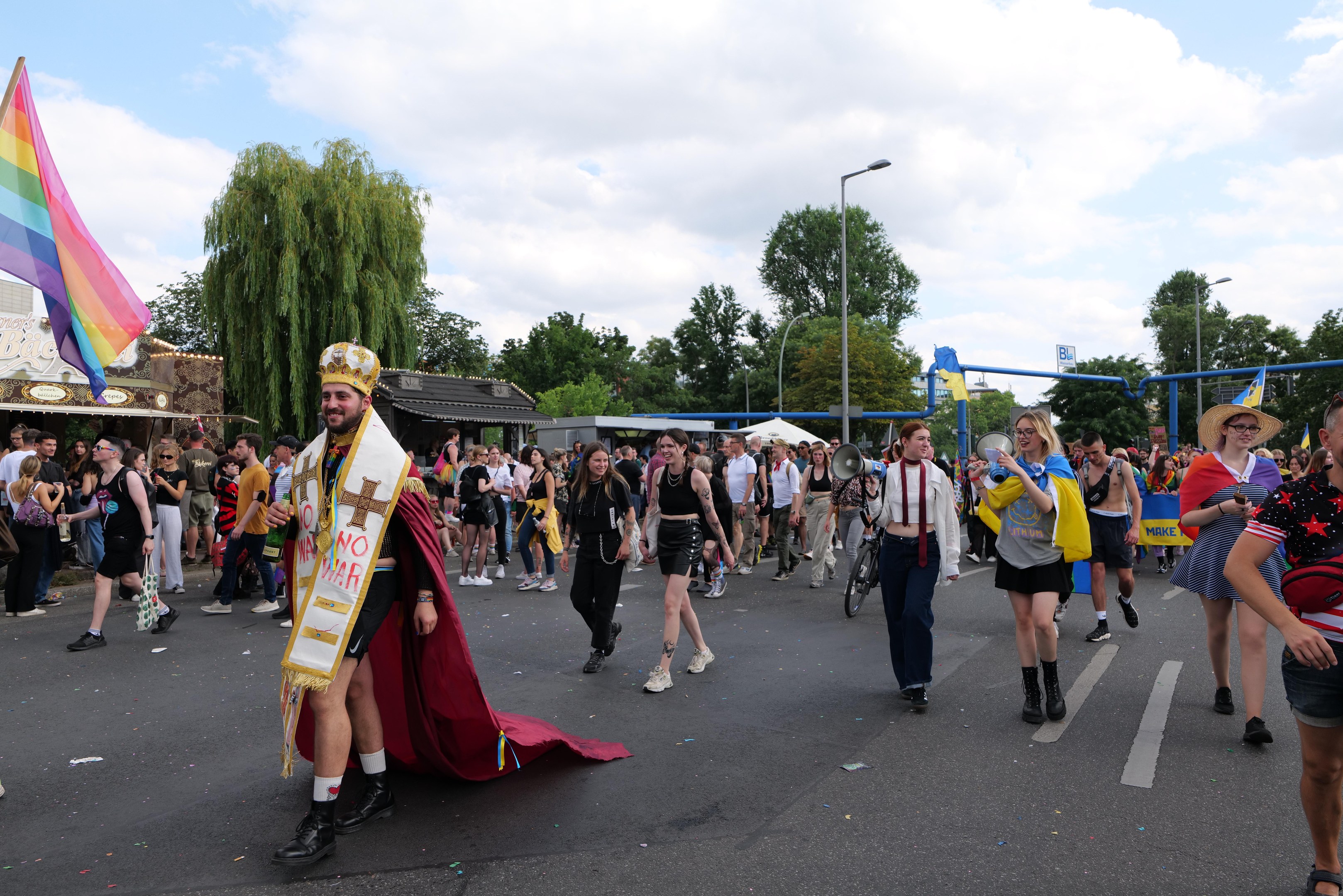 Eine Gruppe von Menschen marschiert bei der Gay Pride Parade 2018, einige mit Musikinstrumenten und Mützen, die eine Regenbogenflagge tragen, auf einer Straße mit Laternenpfählen, Bäumen, Hütten und einem bewölkten Himmel im Hintergrund.