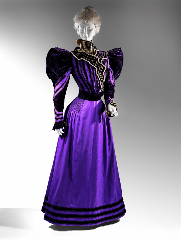 Eine Schaufensterpuppe trägt ein bodenlanges lila Abendkleid mit einem schwarzen und goldenen gemusterten Rock, der mit goldener Stickerei und Perlen verziert ist, eine eng anliegende Taille mit Spitzenbesatz und eine hochgesteckte Frisur mit zarten Ohrringen.