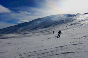 Eine Person mit Helm, Handschuhen und Schuhen fährt auf einer schneebedeckten Piste Ski, hält Ski-Stöcke in den Händen und hat Berge und einen bewölkten Himmel im Hintergrund, während die Sonne auf dem Schnee glänzt.