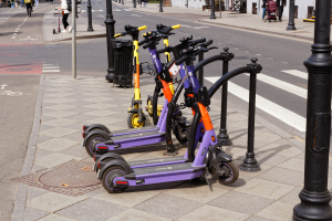 Eine Reihe von E-Scootern, die entlang einer Straße mit Fußgängern und Radfahrern auf dem Gehweg geparkt sind, ein Müllcontainer im Vordergrund und Gebäude, Bäume und Pflanzen im Hintergrund.