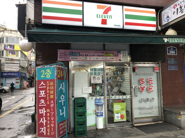 Ein 7-Eleven-Konzern in Seoul, Südkorea, mit Fahrzeugen auf der Straße, einem Fußgänger auf der linken Seite, Gebäuden im Hintergrund und dem Himmel darüber.
