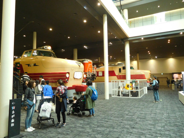 Eine Gruppe von Menschen steht vor einer Bahnausstellung in einem Museum, einige mit Taschen und Kinderwagen, andere betrachten die Ausstellungsstücke, mit Säulen, Geländern, Deckenleuchten und einer Texttafel im Bild.