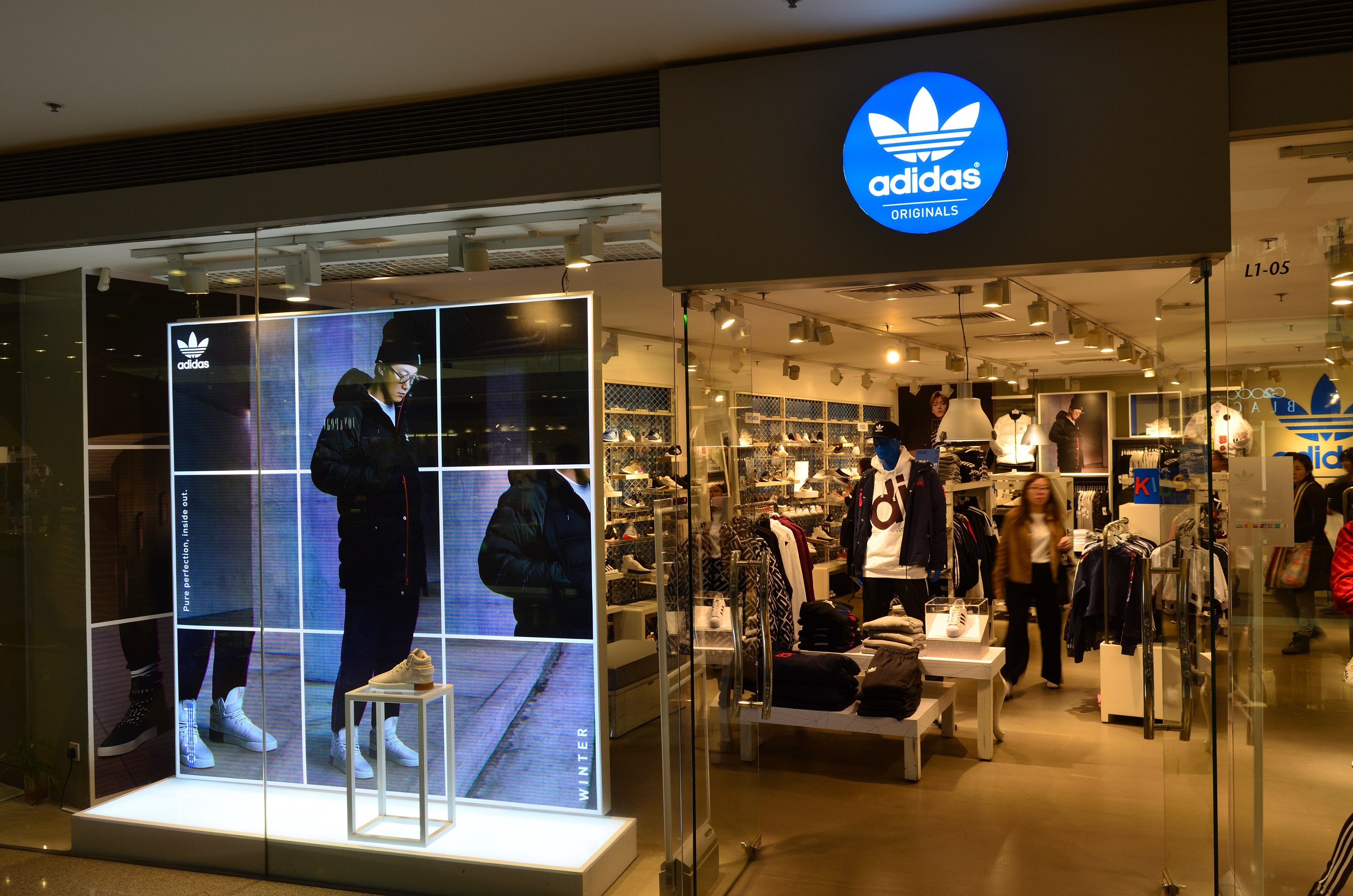 Ein hell erleuchteter adidas Originals-Store in einem Einkaufszentrum, der Puppen, hängende Kleidung, ausgestellte Schuhe und ein großes Adidas-Logo-Board zeigt.