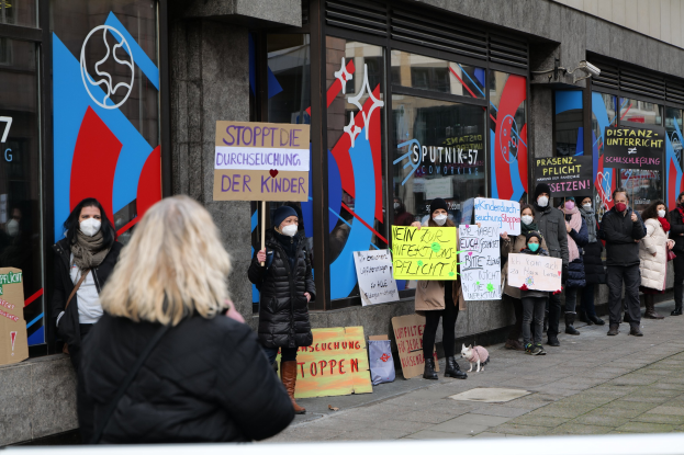 Maskierte Menschen halten Schilder und Plakate vor einem Glasgebäude, mit Kameras und einem Hund, wahrscheinlich protestieren sie gegen ein deutsches Maskenverbot in Schulen.