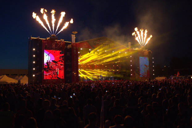 Eine große Menschenmenge auf einem Musikfestival schaut auf der hell erleuchteten Bühne mit Bannern, umgeben von Zelten und Feuerwerk am dunklen Himmel.