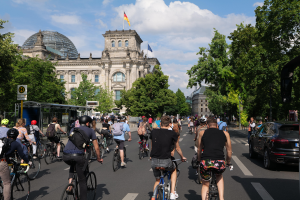 Eine Gruppe von Menschen, die auf Fahrrädern eine von Bäumen gesäumte Straße in Berlin, Deutschland, entlangfährt, mit Gebäuden auf beiden Seiten, einer Bushaltestelle auf der rechten Seite und einer Flagge, die auf einem der Gebäude weht, unter einem bewölkten Himmel.